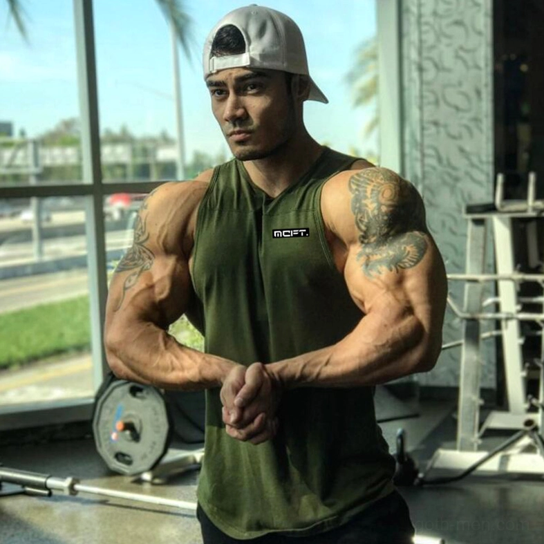 Tattooed flexing man wearing army green gothic tank top with chest logo in gym