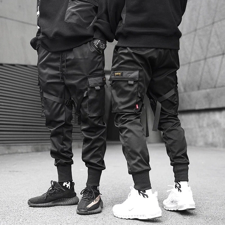 Two male models wearing matching black cargo joggers in outdoor industrial setting demonstrating versatile gothic streetwear styling