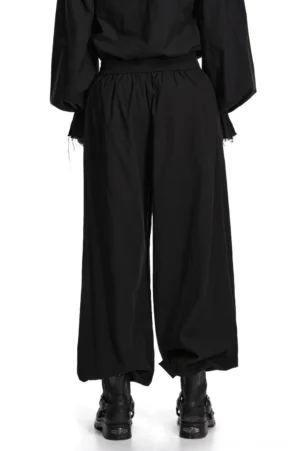 Black gothic pleated lantern tapered pants back view on male model, displaying elastic waistband and graceful drape of gathered ankle cuffs