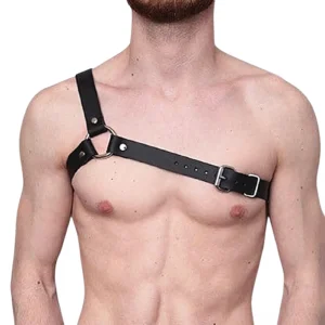 Shirtless male model wearing black leather chest bondage harness with single shoulder strap and metal O-ring, front studio view on white background