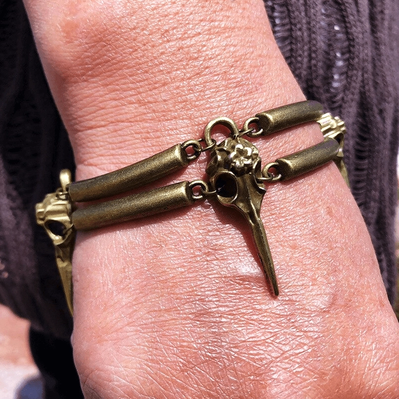Back of hand wearing gothic crow skull bracelet close-up view against dark knitted fabric showing vintage bronze patina