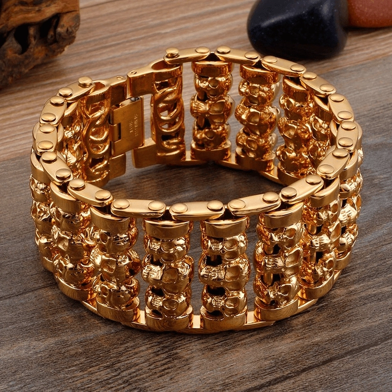 Gold gothic skull chain bracelet with wide band and stacked skull links on stone background