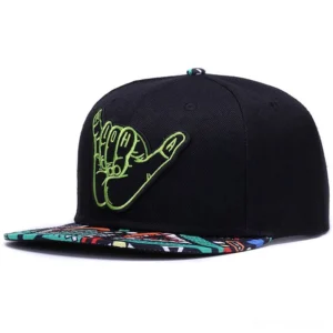 Front three-quarter view of black alternative snapback baseball cap featuring bright green embroidered shaka symbol and patterned brim