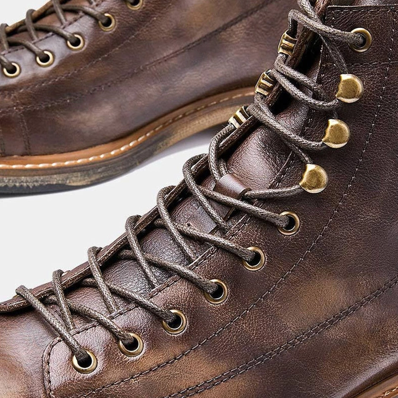 Lace-up detail of gothic men's ankle boots with brass metal hooks and distressed leather texture frontal close-up