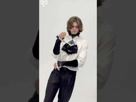Male goth model wearing black velvet gothic arm gauntlets as part of complete formal goth outfit including high-waisted trousers and matching accessories, three-quarter studio shot with gray background