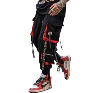 Black gothic cargo joggers with multi-pocket straps worn by tattooed male model, side view showing red-black strap accents and decorative details