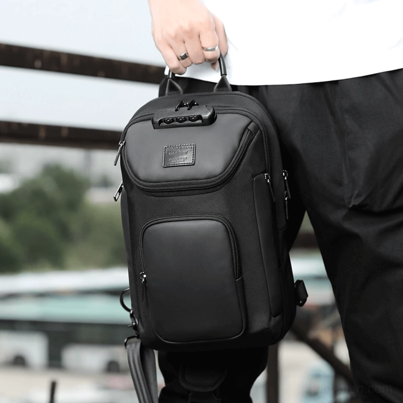 Male model holding black sling crossbody bag with combination lock low front angle urban background