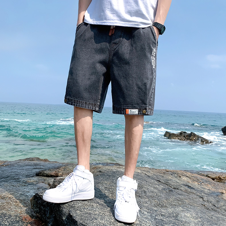 Male model wearing dark grey gothic denim shorts with white sneakers at coastal beach setting, front view