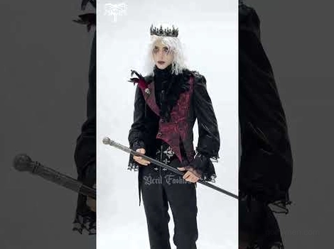 Male model styled in complete gothic formal outfit with wine red and black waistcoat, crown, decorative waist chain, and cane against dark background