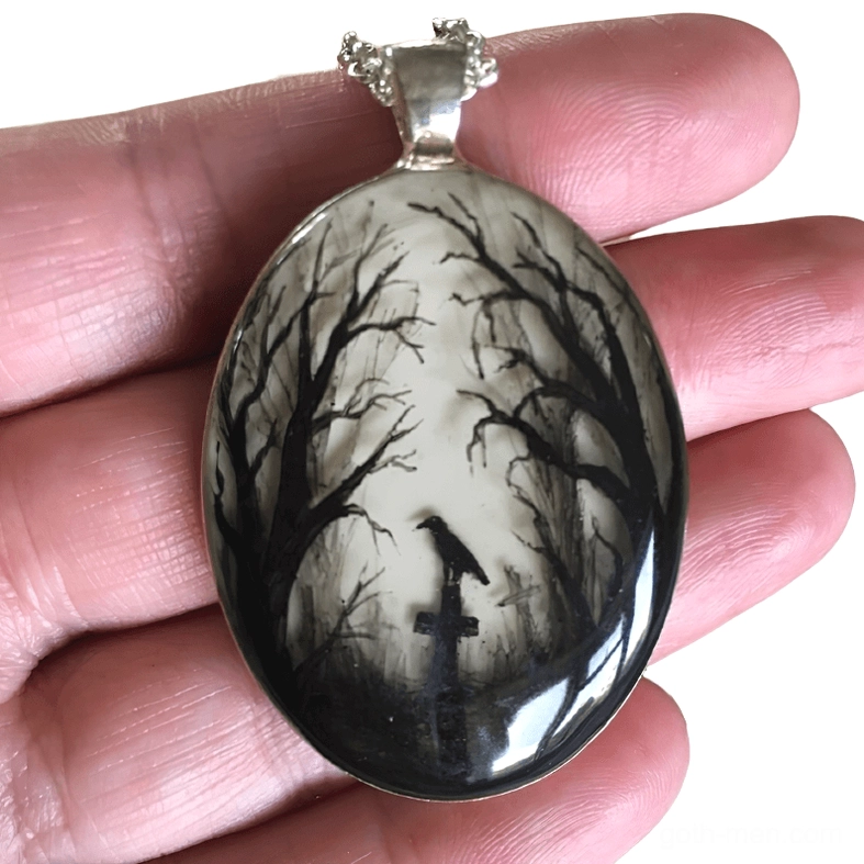 Close-up detail view of gothic graveyard pendant held in hand, highlighting intricate hand-painted black and white raven and tombstone design with silver bail
