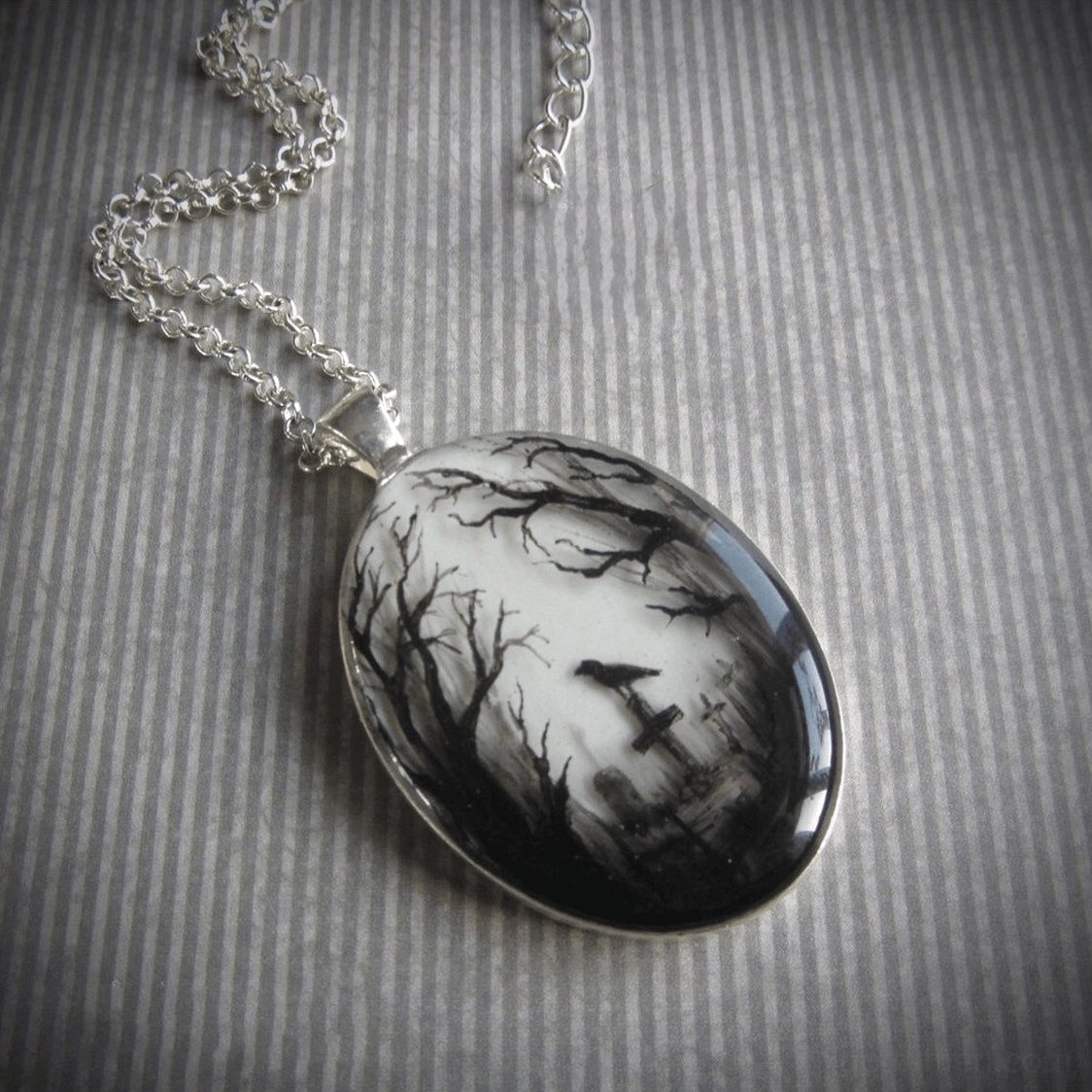 Gothic graveyard hand-painted pendant necklace draped on fabric surface, displaying silver link chain and monochromatic occult raven motif