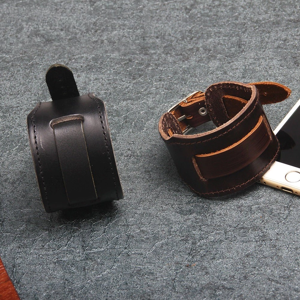 Two wide leather gothic cuff bracelets in black and brown displayed on dark desktop surface with smartphone accessory