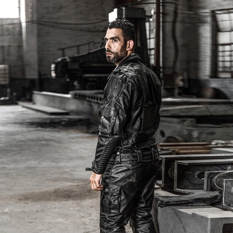 Male model wearing black genuine leather gothic motorcycle jacket, left side standing view, detailed stitching visible, metal hardware details