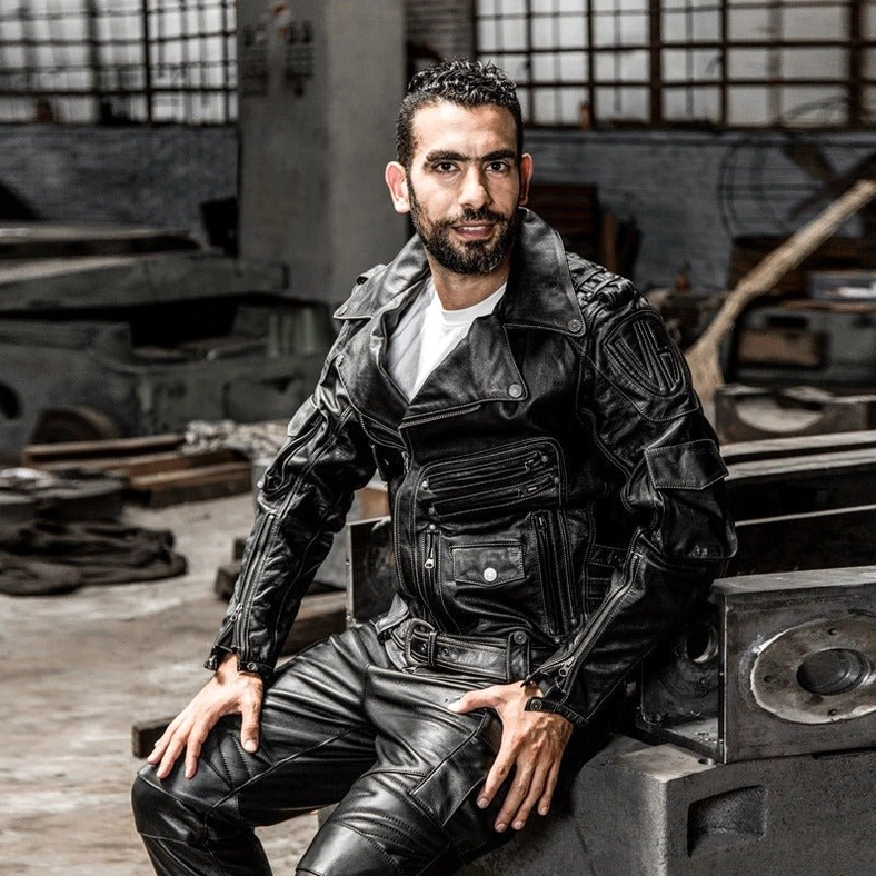 Male model wearing black genuine leather gothic motorcycle jacket, fitted rider silhouette, industrial workshop setting, full outfit styling
