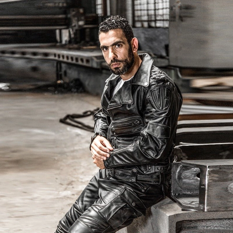 Male model wearing black genuine leather gothic motorcycle jacket and leather pants, relaxed sitting pose, industrial aesthetic, complete biker outfit