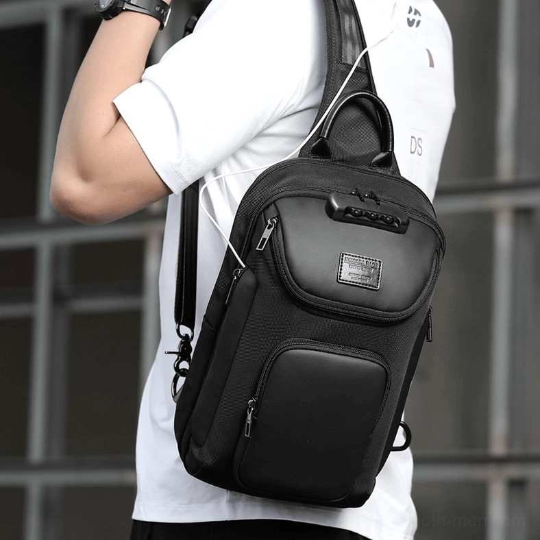 Male model wearing black sling crossbody bag on left shoulder with headphone routing hole urban building background