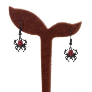 Mini black spider dangle earrings displayed on brown velvet ear stand, showing crimson crystal accents, gothic accessories close-up