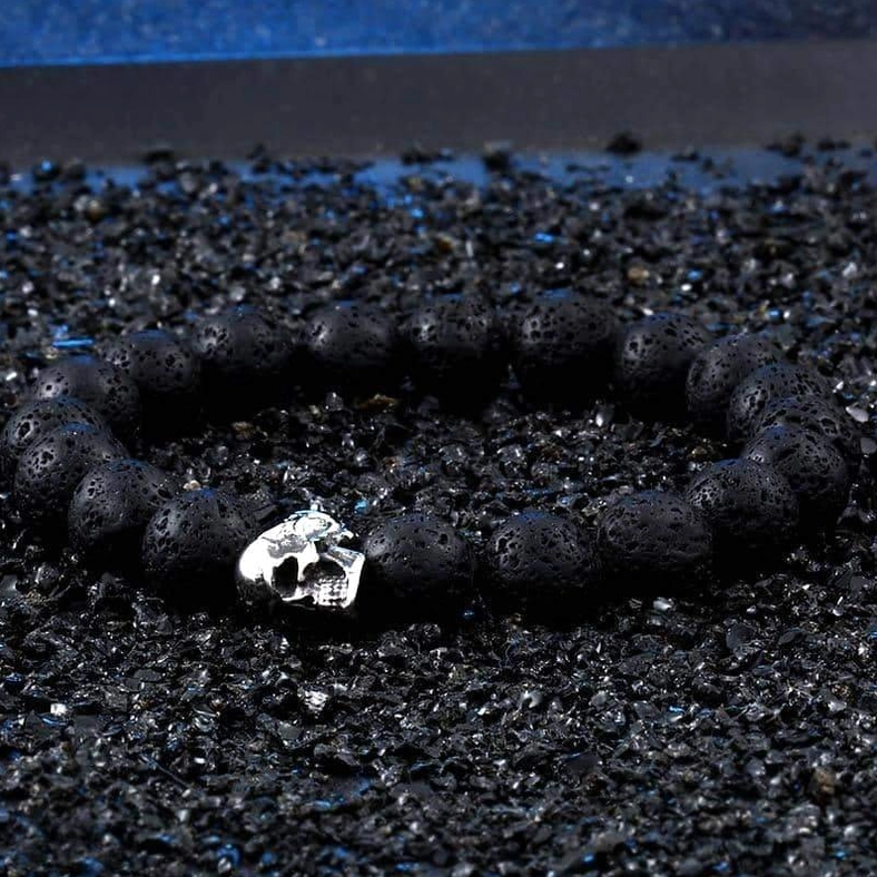 Black porous lava stone beads with silver skull charm on black gravel surface