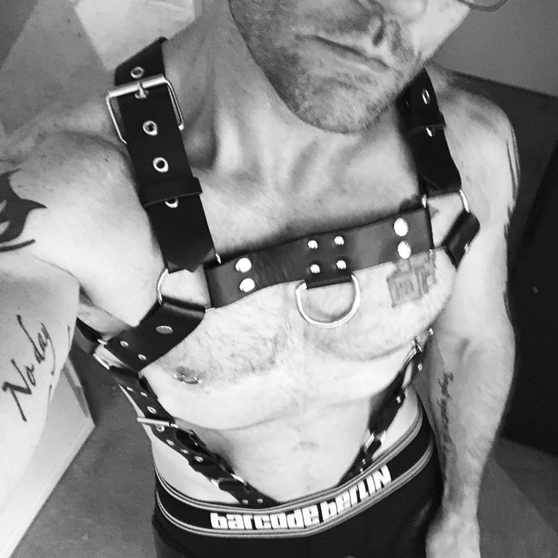 Man wearing black metal rivet leather chest harness with central O-ring, tattooed shirtless model showcasing gothic bondage harness fit