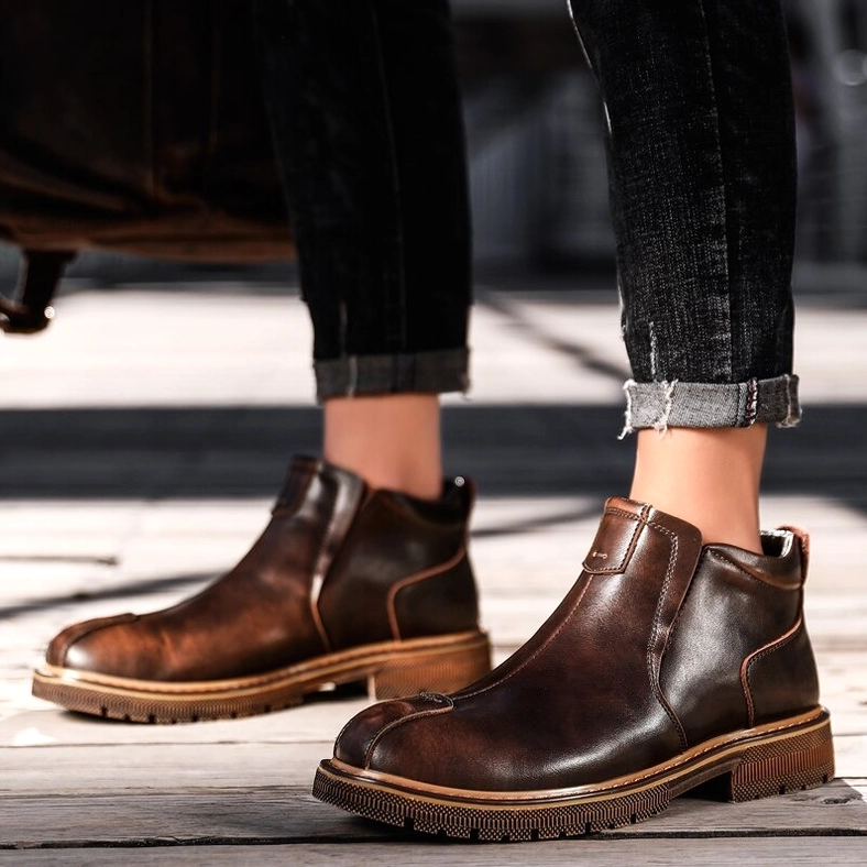 Man wearing brown gothic Chelsea boots with rolled dark jeans on wooden floor, front-facing casual stance