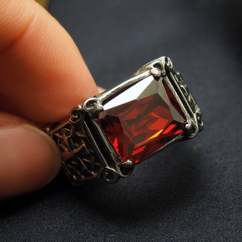 Red gemstone celtic cross gothic ring worn on finger, close-up side view displaying faceted stone and engraved cross patterns against dark fabric