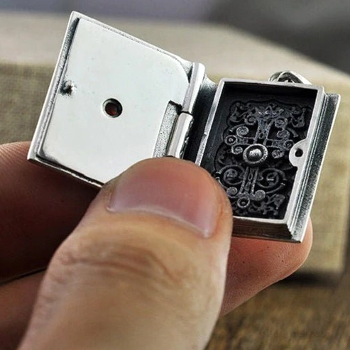Openable silver bible locket pendant in human hand showing compact size and intricate interior cross carving, displayed on wooden surface
