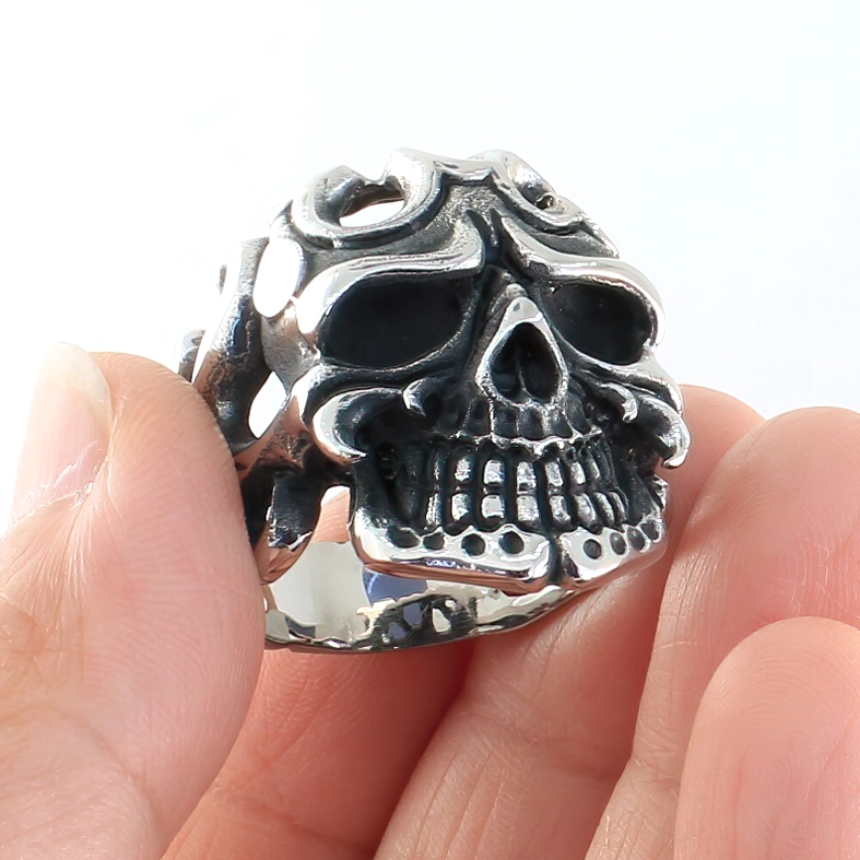 Close-up side view of silver gothic skull ring held between fingertips, displaying intricate hollow tribal design and polished details