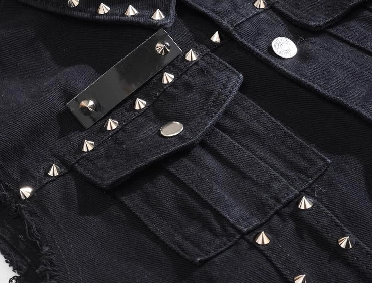 Angled close-up of black denim vest pocket detail with spike studs and fasteners