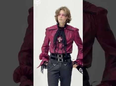 Male model wearing wine red Victorian gothic shirt with cross pendant decoration against dark studio background, styled with belted pants